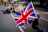 An elderly woman sits down on a chair in the middle of the road while holding Union flag.