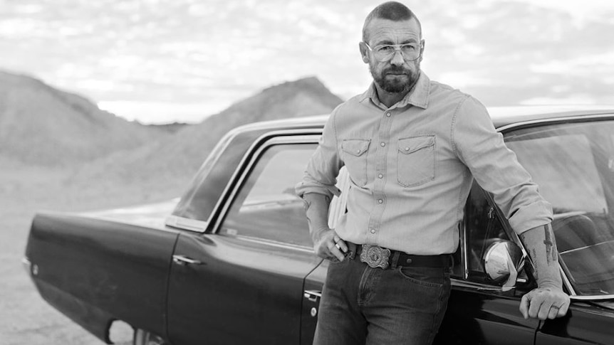 balck and white photo of man in spectacles with short hair and beard leaning on a car