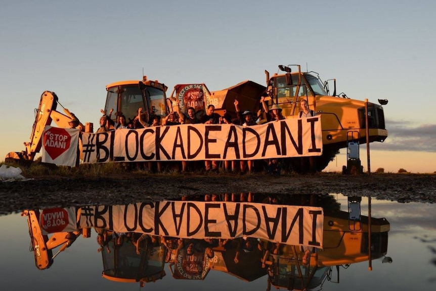 Protesters stand behind a banner saying Blockade Adani in front of construction equipment.
