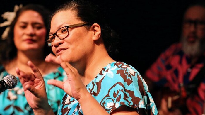Photo of Rita Seumanutafa wearing a blue dress with flowers. She is holding her hands up and there is a mic infront of her.