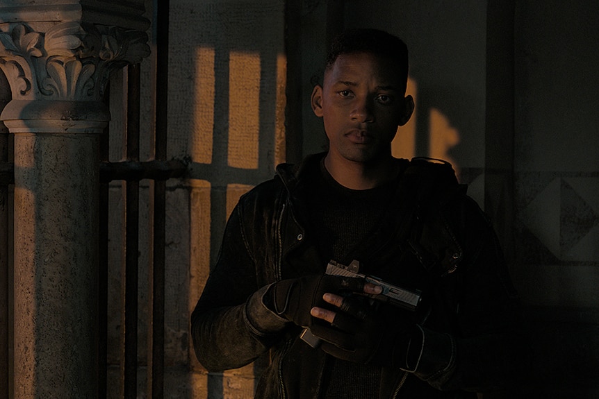 Will Smith stands in the shadows wearing a black outfit and holding a pistol to his chest.
