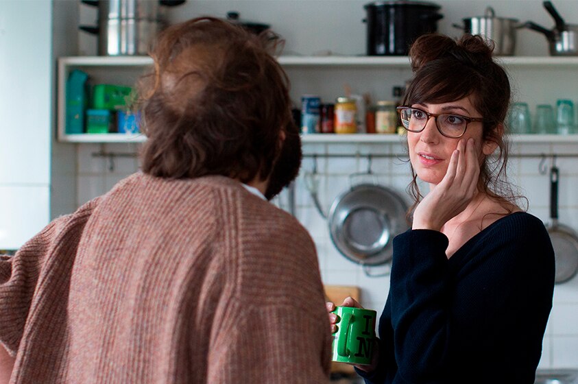 A brown haired man and woman with glasses stand and speak to each other in natural light filled kitchen with pots and pans.