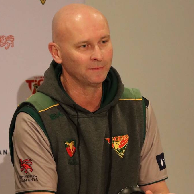 A bald man wearing a green cricket Tasmania jumper with a tiger logo on it stares into the distance.
