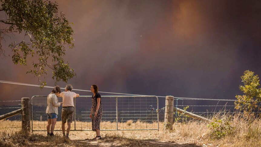 Three people stand at a fence with black, smoky skies in the background.