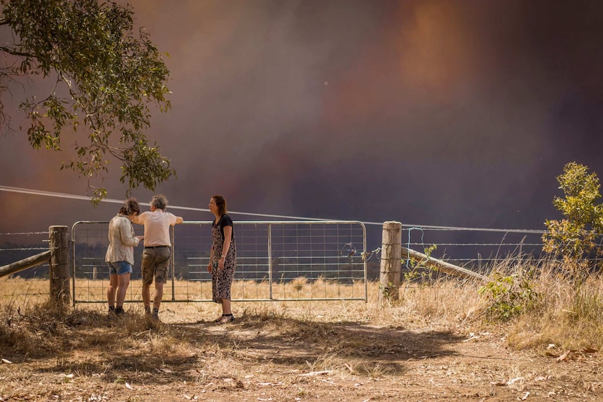 Three people stand at a fence with black, smoky skies in the background.