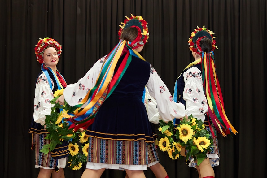 Three women wearing colourful headpieces dance on a stage holding hands.