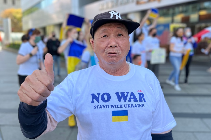 An older man wearing a NO WAR T-shirt with a Ukrainian flag gives a thumbs up to the camera