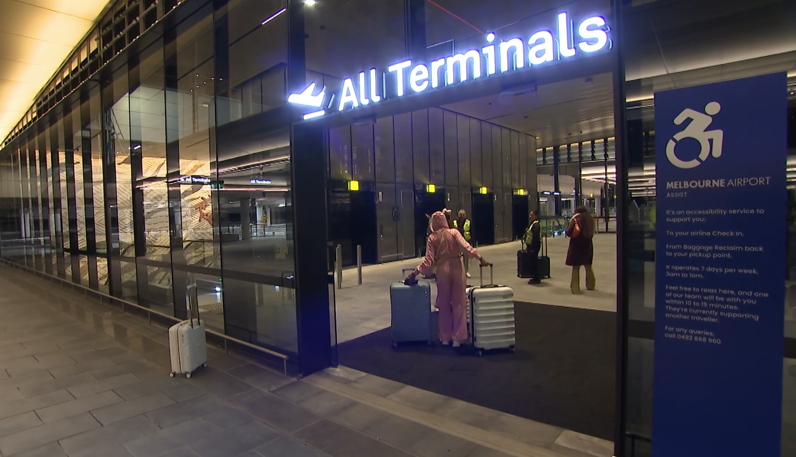 A traveller dressed in a pink tracksuit wheeling a white suitcase follows others through a door that says "all terminals".