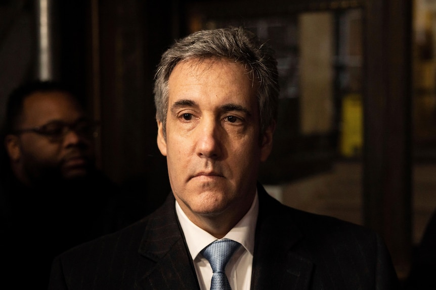 a close up Michael Cohen wearing a black suit with a pale blue tie. He has a serious expression on his face.
