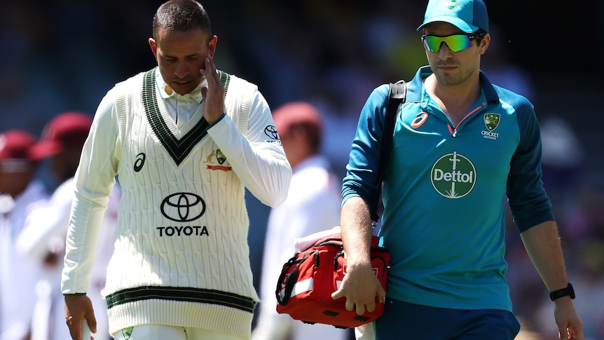 Usman Khawaja looks down and touches the side of his face as he walks off the field with a team doctor