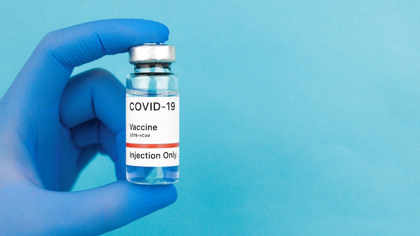 Gloved hand holding vial of clear liquid with a label saying COVID-19 vaccine, in story about what to expect getting vaccine.