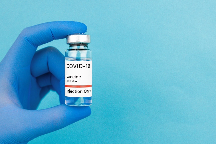 A hand seen with a blue plastic glove on it against blue holds a vial of clear liquid with a label saying COVID-19 vaccine