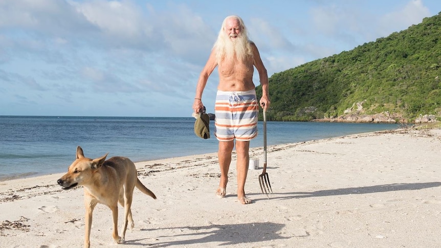 A man with long white hair and beard, wearing red and blue striped baordshorts, walks on sand near sea, next to a walking dingo.