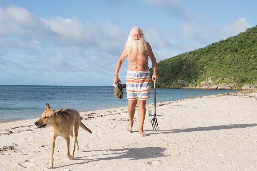 A man with long white hair and beard, wearing red and blue striped baordshorts, walks on sand near sea, next to a walking dingo.