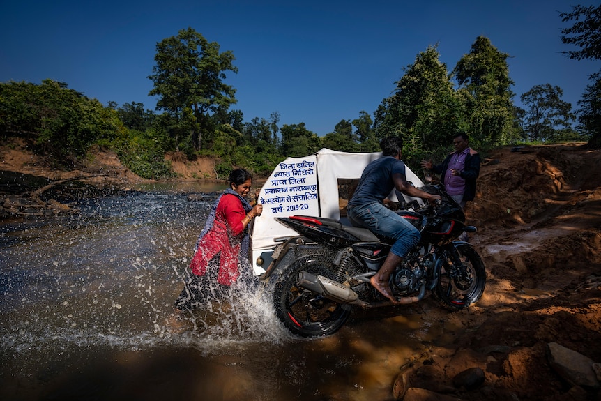 A woman pushes an ambulance sidecar attached to a motorbike with a man on it, up a riverbank.
