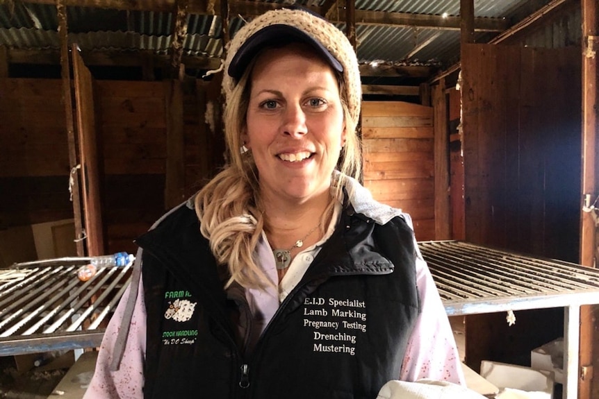 A blonde woman in a black vest, cap and beanie smiles, standing in a shearing shed.