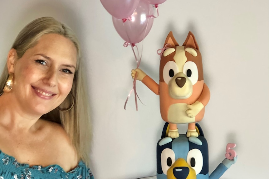 Woman smiling beside an extravagant 5th birthday cake featuring the ABC Kids TV characters Bluey and Bingo.