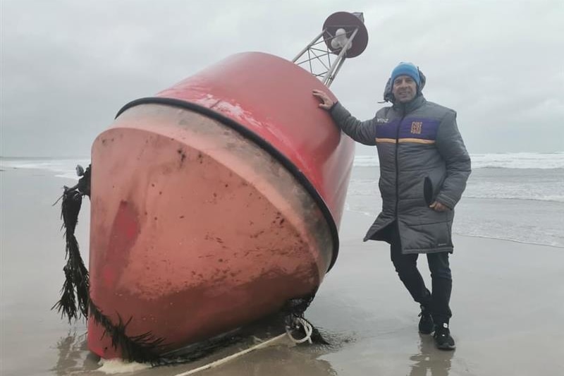 Man stands on beach leaning against red buoy the size of a small car