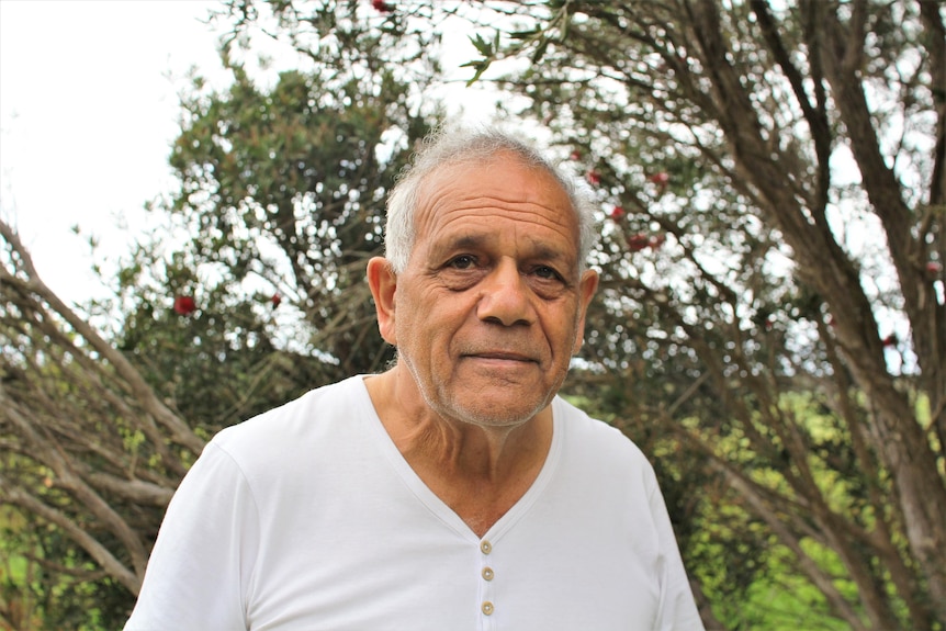A dark skinned man with white hair and a white shirt on looks at the camera with a solemn expression 
