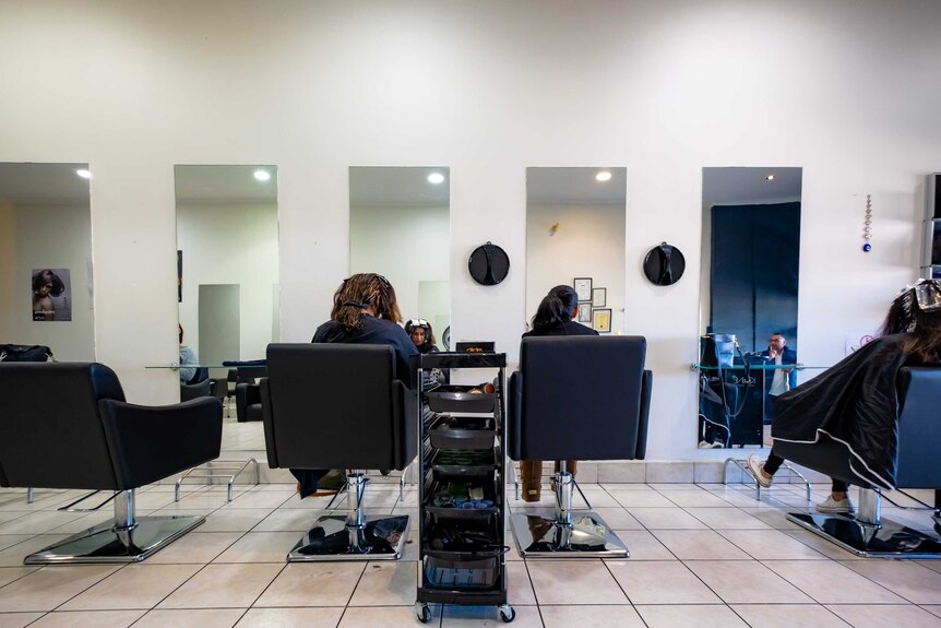 You view women sitting in black salon chairs from behind as long portrait-size mirrors show you their faces looking down.