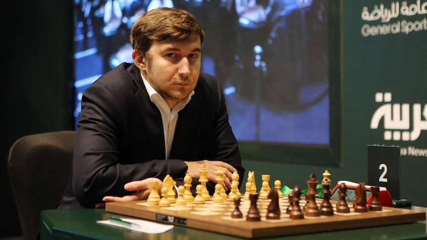A man in a jacket looks at the camera in front of a chess board