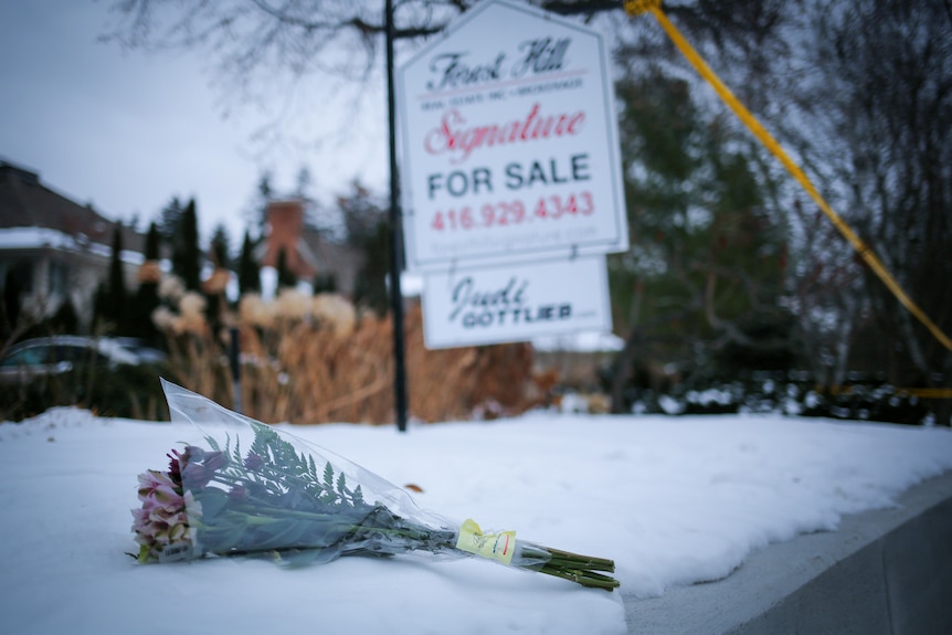 A bunch of flowers left on the snowy ground next to a For Sale sign