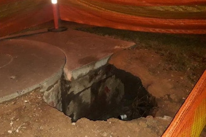 A large, jagged hole in a concreted area with hazard fencing around it.