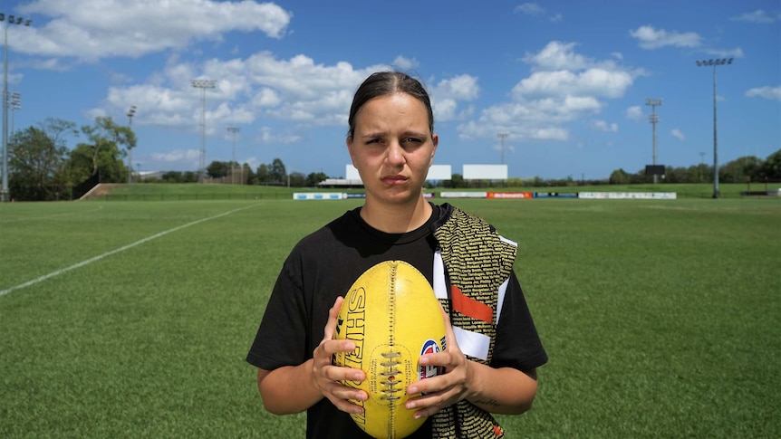 Danielle Ponter holds a football and sports an NT Thunder jersey slung over her shoulder in front of a lush playing field.