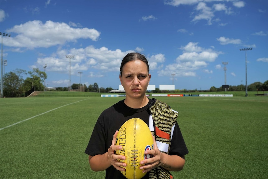 Danielle Ponter holds a football and sports an NT Thunder jersey slung over her shoulder in front of a lush playing field.