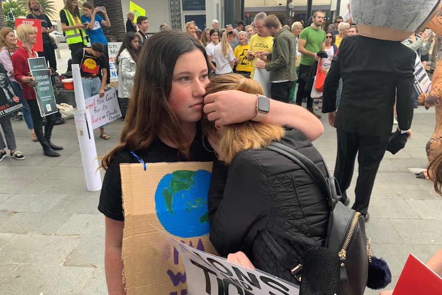 a girl comforts another girl both wearing black with signs hung around their necks