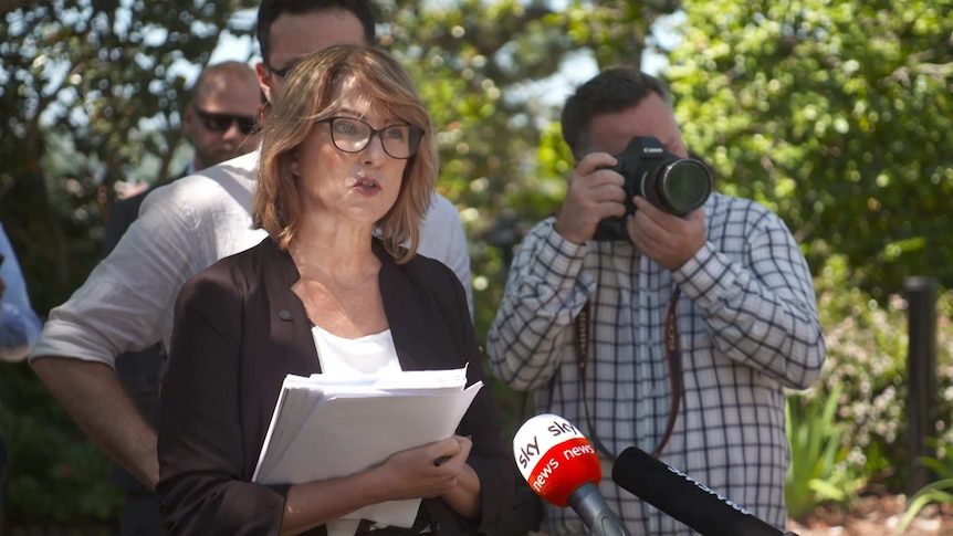 Woman holding notepad standing in front of microphones next to a photographer.