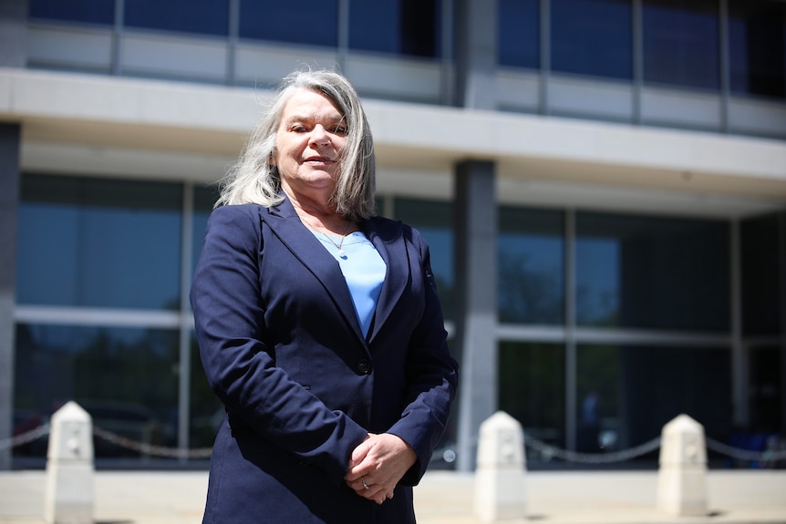 A woman wearing a navy blue jacket and grey hair stands in front of parliament