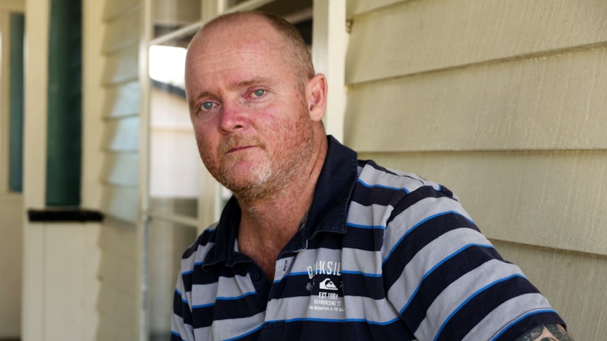 Aman in a striped polo shirt sits on a front porch and looks at the camera with a sombre expression.