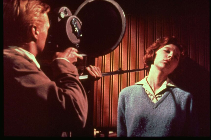 A still from the 1960 film Peeping Tom with a man holding a camera and filming a woman leaning against a wall