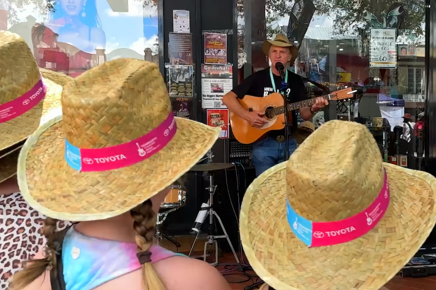 A man with a guitar performs in front of three girls in hats
