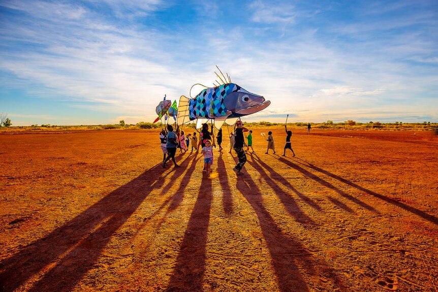 A colourful big fish walks across the sunset lit red dirt surrounded by children.