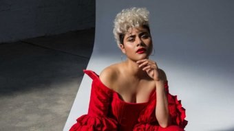 Montaigne wears a red dress and black shoes with fishnet socks. She bends down in front of white background and looks to camera.