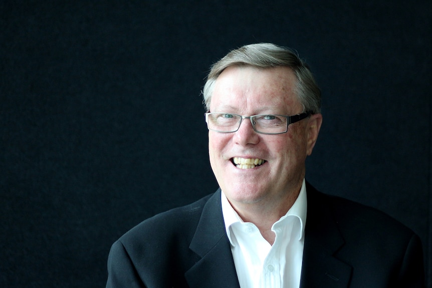 Alan Behm, a man wearing spectacles, a white shirt and dark blazer, poses for a photo, smiling