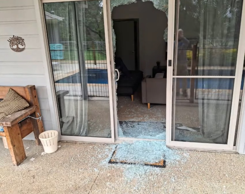 Broken glass covers a door mat and the ground outside a sliding glass door that has been fully smashed.