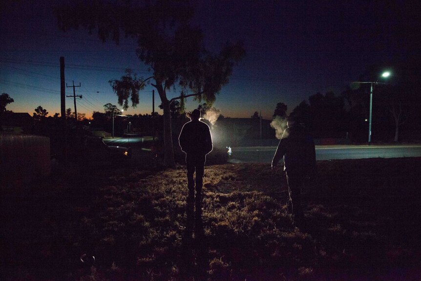 The silhouette of two men in cowboy hats standing in pre-dawn light on the grassed area of a town park.