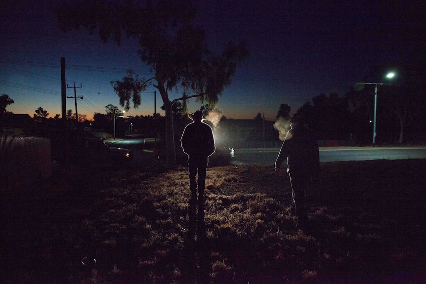 The silhouette of two men in cowboy hats standing in pre-dawn light on the grassed area of a town park.
