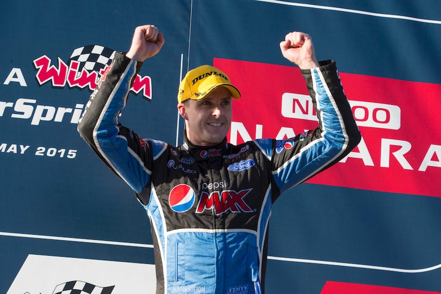 A man holds his arms up in a victory gesture, fist clenched, wears yellow, cap, black and blue racing gear, banner behind.