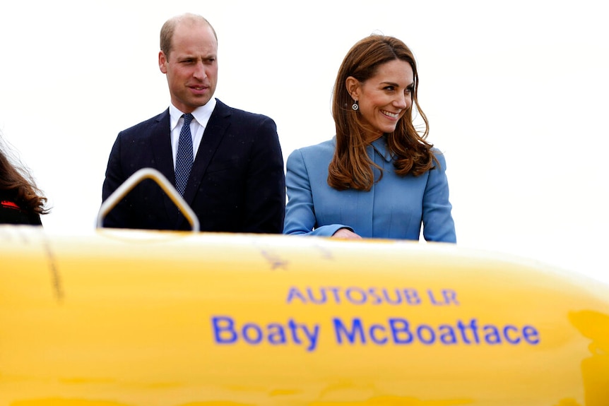 William and Kate stand behind part of a yellow submarine. Will's expression is quizzical while Kate is smiling.