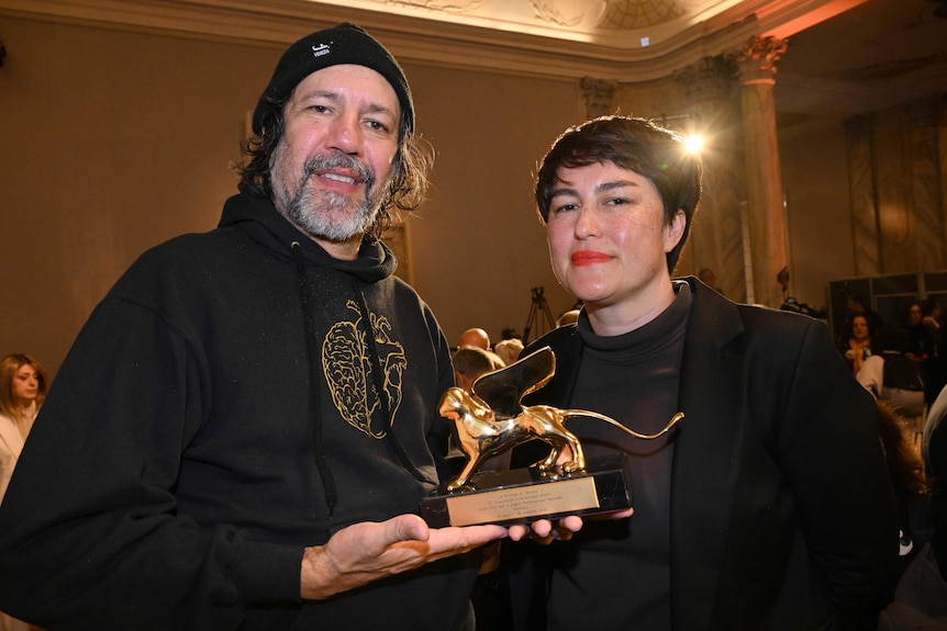 A Bigambul and Kamilaroi artist wearing a black hoodie holds a golden lion trophy with an Asian Australian woman with dark hair.