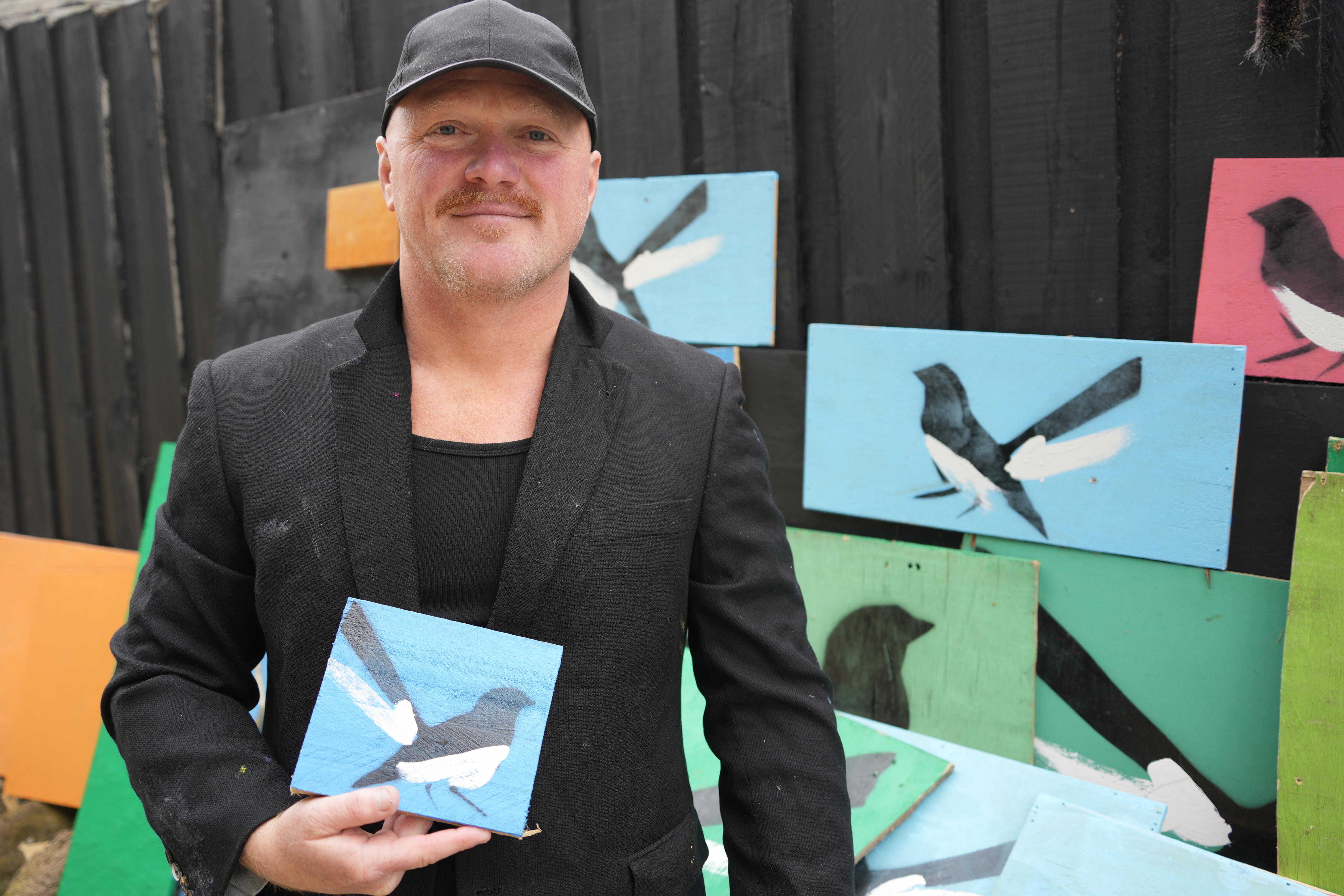 Mike Mclean holds up a willie wagtail painting with other similar paintings against a fence behind him.