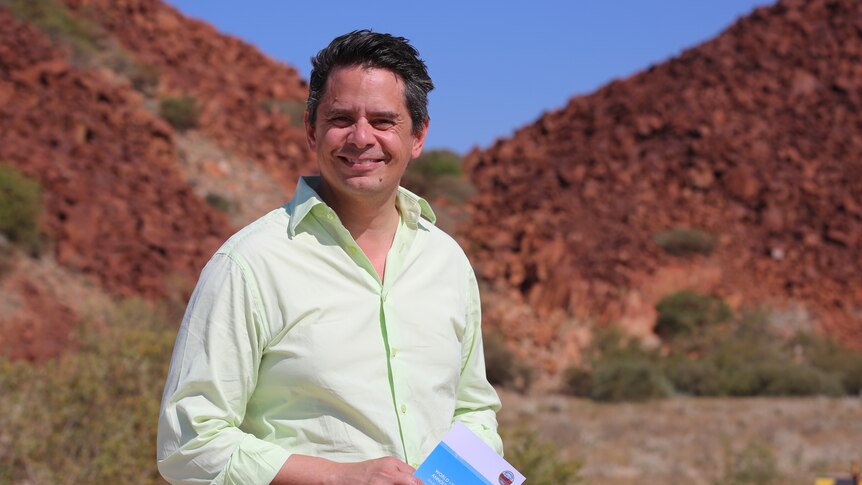 A man in a business shirt stands smiling in the foreground of a rocky red landscape
