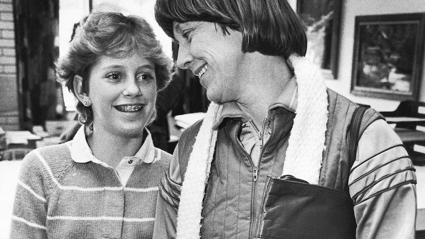 A black and white photo of a teenage girl with braces smiling as her mother looks down at her. 