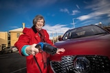 A woman wearing a red coat holds a car charging plug, smiling. She's standing next to a red MG.