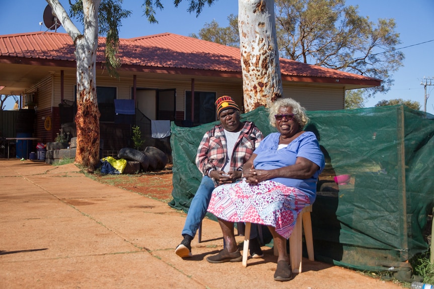 Two elderly Aboriginal women sit in front of a house.