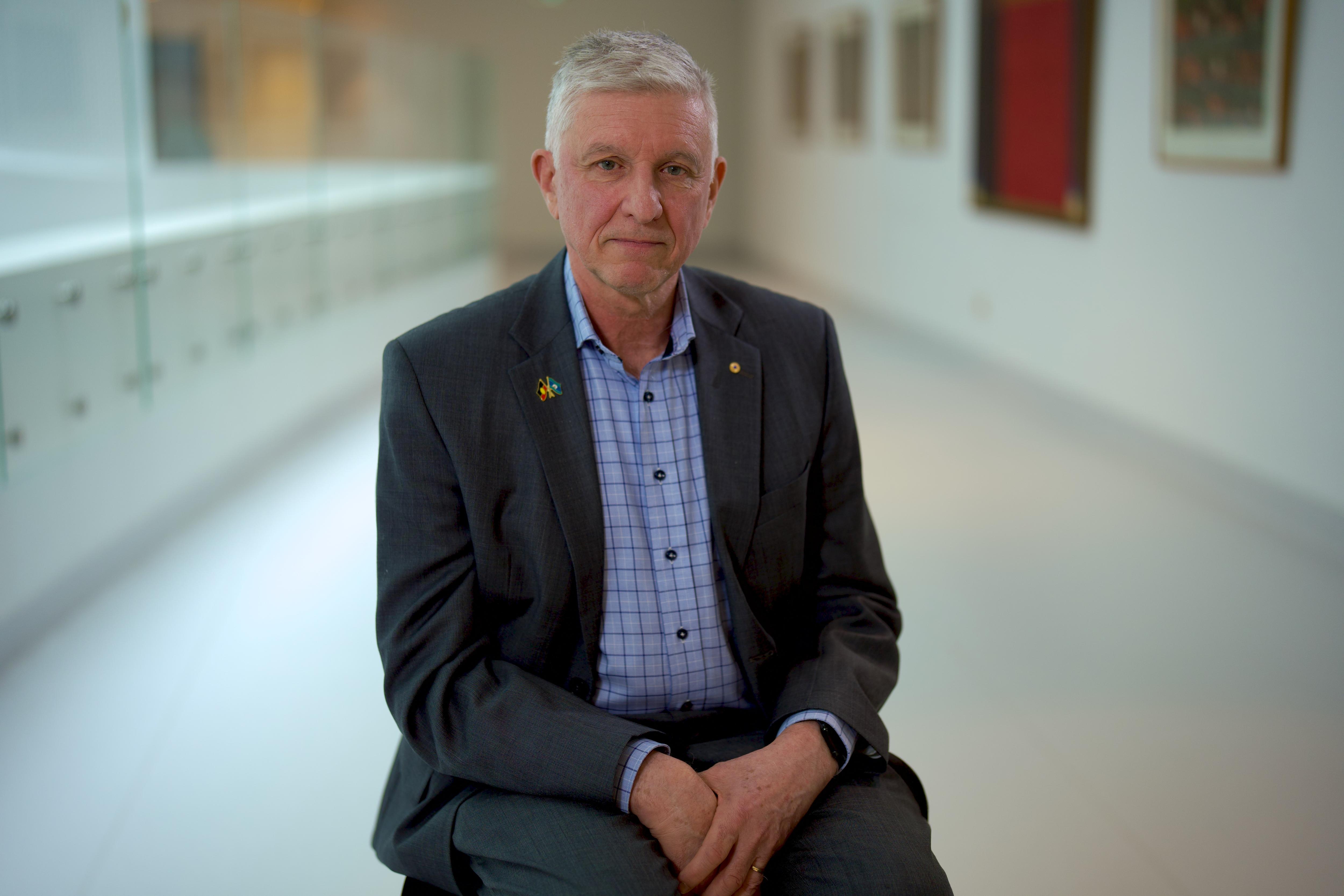 Grant McArthur sitting in a chair in a hospital hallway. Wearing a blue check shirt and suit